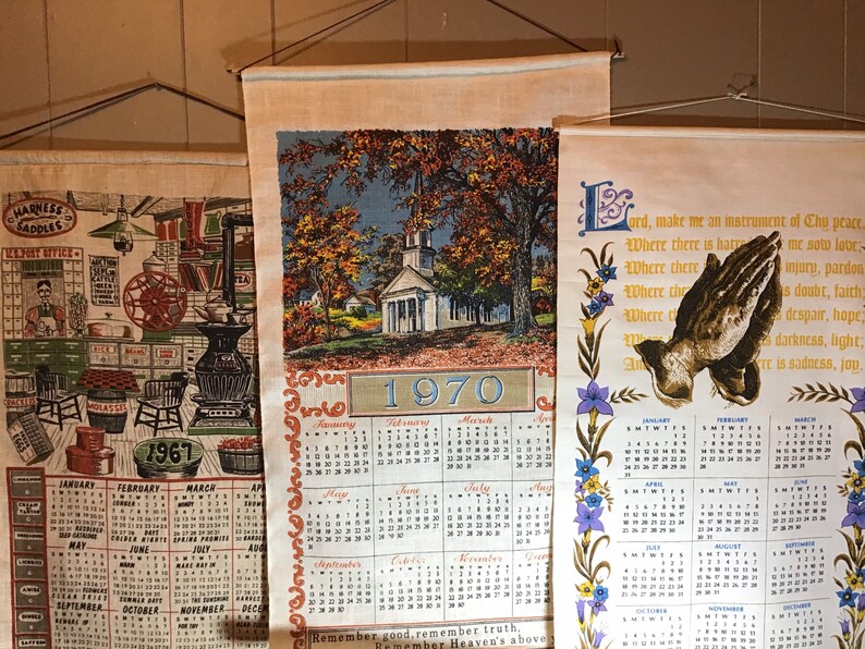 May include: Three vintage wall calendars with a country theme. The first calendar is from 1967 and features a black and white illustration of a general store. The second calendar is from 1970 and features a colorful illustration of a white church with a steeple. The third calendar features a prayer with a dove and a floral border. All three calendars have a cream background and are printed with black text.