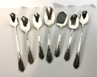 May include: Set of seven silver spoons with ornate handles. The spoons have a polished finish and are arranged in a row on a white surface.