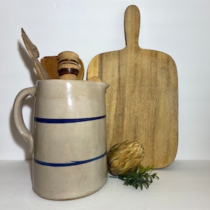 May include: A vintage, blue and white, stoneware pitcher with a handle. The pitcher is sitting on a wooden cutting board with a wooden spoon and a wooden utensil.