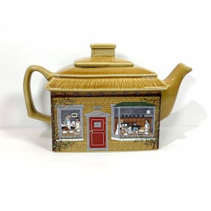 May include: A tan ceramic teapot shaped like a house with a thatched roof. The house-shaped body features a brick facade with windows and a red door. The teapot has a handle, spout, and a chimney.