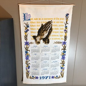May include: A white fabric wall hanging with a prayer printed in a vintage font. The prayer is surrounded by a floral border with purple, yellow, and blue flowers. The hanging also features a calendar for the year 1971 with a picture of praying hands.