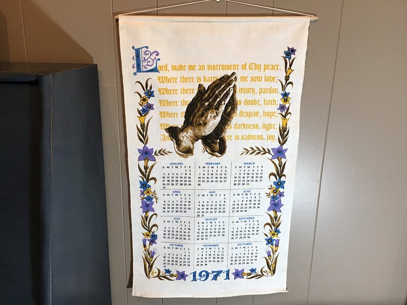 May include: A white fabric wall hanging with a prayer printed in a vintage font. The prayer is surrounded by a floral border with purple, yellow, and blue flowers. The hanging also features a calendar for the year 1971 with a picture of praying hands.