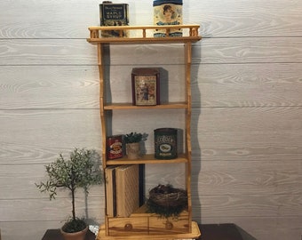 May include: A wooden shelf unit with three shelves. The top shelf has two vintage tin cans, one with a picture of a woman and the text "Maple Syrup" and the other with a picture of a woman. The middle shelf has a vintage tin can with a picture of a woman and the text "Hershey's Kisses". The bottom shelf has a vintage tin can with the text "Golden", a small potted plant, and a stack of books.