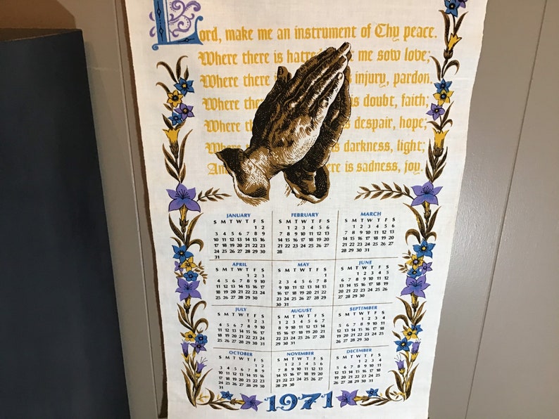 May include: A vintage kitchen towel with a floral border and a calendar for the year 1971. The towel features a prayer in a decorative font and an image of praying hands.