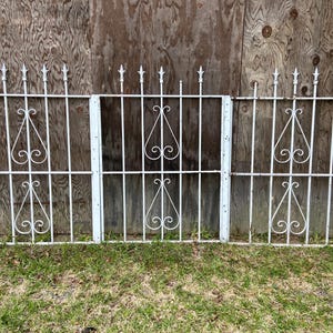 May include: Three white metal gates with decorative scrollwork and pointed finials. The gates are set against a wood background.