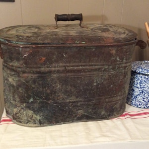 May include: An antique copper wash tub with a wooden handle on the lid. The tub has a patina of age and is oval shaped.
