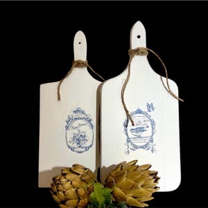 May include: Two white wooden cutting boards with blue and white designs. The board on the left has a floral design with the text "Farm Flowers" and the board on the right has a bird design with the text "From Our Kitchen".