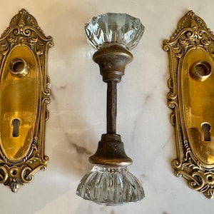 May include: Antique door hardware set featuring a clear glass door knob with a faceted design and a brass-toned metal stem. Two ornate brass door plates with keyholes flank the knob, showcasing a vintage aesthetic.
