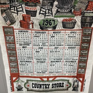 May include: A vintage 1967 calendar tea towel with a country store theme. The towel features a calendar with each month's name and days of the week. The calendar is surrounded by illustrations of various food items, including coffee, molasses, pickles, and eggs. The towel also features a red star with the words "Country Store" written below it.