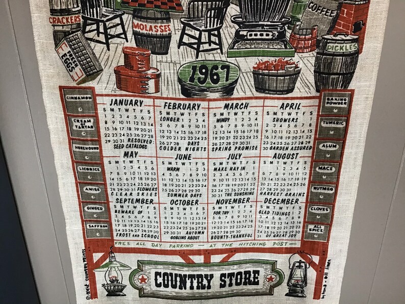 May include: A vintage 1967 calendar tea towel with a country store theme. The towel features a calendar with each month's name and days of the week. The calendar is surrounded by illustrations of various food items, including coffee, molasses, pickles, and eggs. The towel also features a red star with the words "Country Store" written below it.
