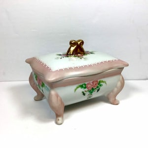May include: A white porcelain trinket box with a pink and gold trim. The box has a floral design with pink roses and green leaves. The lid has a gold knob in the shape of a knot.