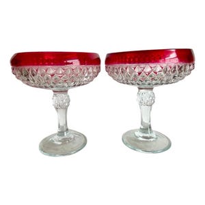 May include: Two clear glass pedestal bowls with a red rim. The bowls have a diamond pattern and are on a white background.