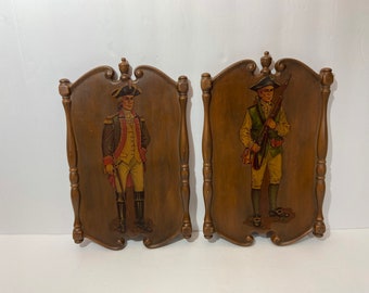 May include: Two wooden plaques with painted images of men in military uniforms. The plaques are brown with a decorative border. The men are wearing tri-cornered hats and holding muskets.