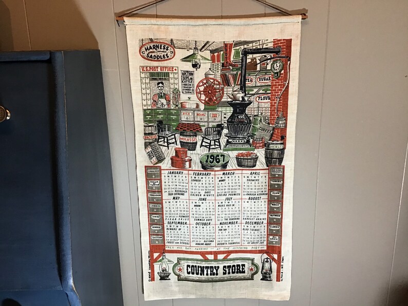 May include: A vintage 1967 calendar tea towel featuring a country store scene with a red, white, and blue color scheme. The towel includes a calendar for each month of the year and the text "Harness Saddles", "U.S. Post Office", and "Country Store".