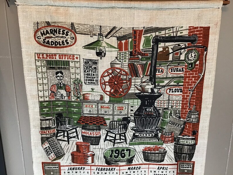 May include: A vintage 1967 calendar tea towel with a red, white, and green color scheme. The towel features a depiction of a general store with various items for sale, including boots, sugar, flour, coffee, and pickles. The text "HARNESS SADDLES" and "U.S. POST OFFICE" are visible at the top of the towel.