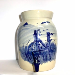 May include: A ceramic water dispenser with a blue and white painted scene of a fence and a birdhouse in a field. The dispenser has a blue spigot.