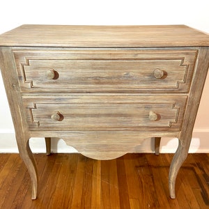 May include: A two-drawer wooden chest with a distressed, light-brown finish. The chest features decorative drawer fronts and round knobs. The legs are curved, and the overall design is vintage-inspired.