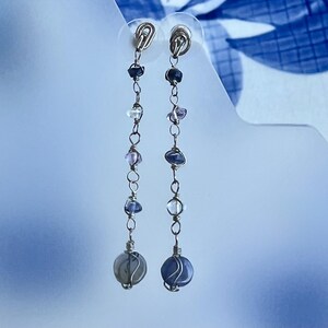 Peut inclure: Paire de boucles d'oreilles pendantes avec un design délicat. Elles présentent une série de petites pierres précieuses colorées enfilées sur un fil argenté. Les pierres sont un mélange de violet, de bleu et de transparent, avec une pierre grise ronde plus grande en bas.