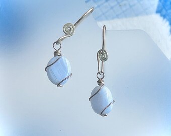 May include: Dangle earrings with oval, pale blue gemstones wrapped in copper wire. The silver-toned ear wires have a spiral design. The earrings are set against a soft, light blue background.