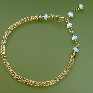Pode incluir: Uma pulseira de malha dourada com um design delicado. A pulseira apresenta uma série de pequenas ligações entrelaçadas, com um fecho e uma fileira de pequenas contas azuis claras. A pulseira está sobre um fundo verde.
