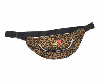 May include: A leopard print fanny pack with a black adjustable strap and buckle. The bag features a white zipper and a small embroidered red rose with green leaves. The fanny pack is a stylish accessory.
