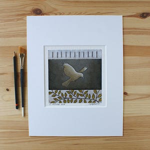 May include: A square print in a white mat featuring a central dark gray panel with a light-colored bird in flight. The top section has vertical lines, and the bottom shows gold-toned leaves. The text 17 octobre 2018 is visible at the bottom left.