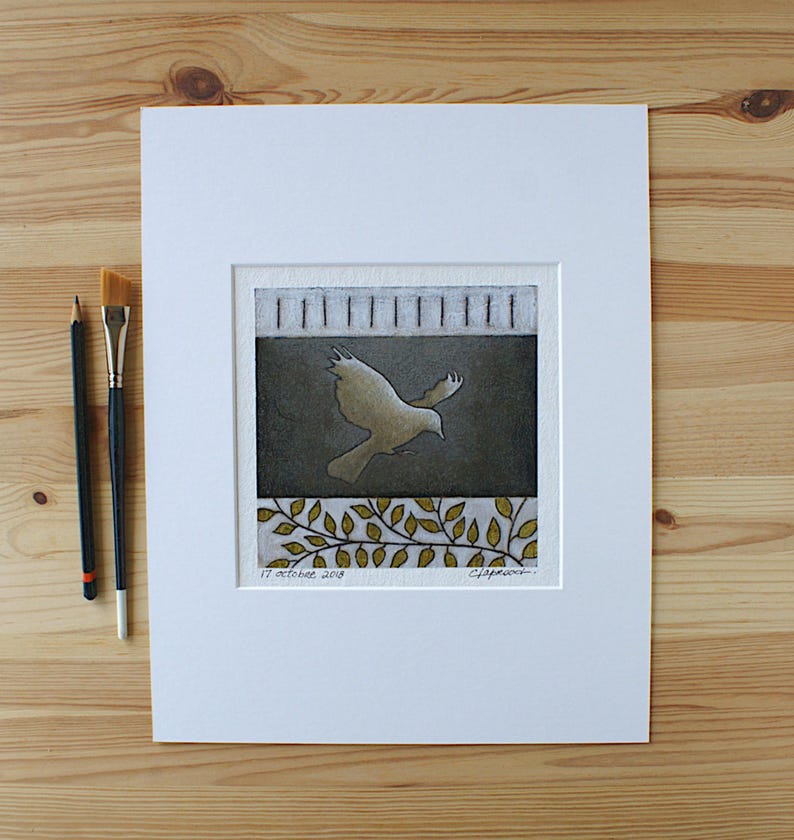 May include: A square print in a white mat featuring a central dark gray panel with a light-colored bird in flight. The top section has vertical lines, and the bottom shows gold-toned leaves. The text 17 octobre 2018 is visible at the bottom left.