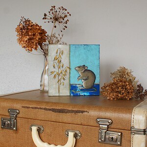 May include: A small painting on a canvas with a blue and white background. The painting depicts a brown mouse sitting on a blue surface with a stalk of wheat in the background.