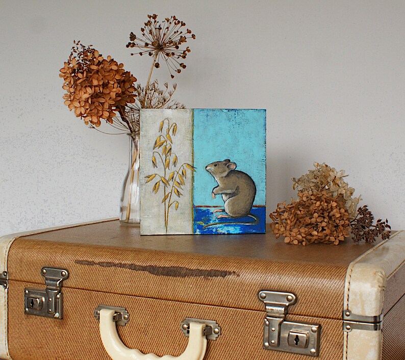 May include: A small painting on a canvas with a blue and white background. The painting depicts a brown mouse sitting on a blue surface with a stalk of wheat in the background.