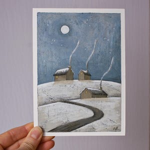 May include: A rectangular art print featuring a winter landscape painting with three small houses on a snow-covered hill under a dark blue night sky. A full moon and stars are visible above, while thin trails of smoke rise from each chimney.