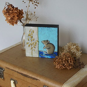 May include: A small painting on a wooden panel depicts a brown mouse standing on a blue and green background. The mouse is looking to the left, and there is a stalk of oats on the left side of the painting.