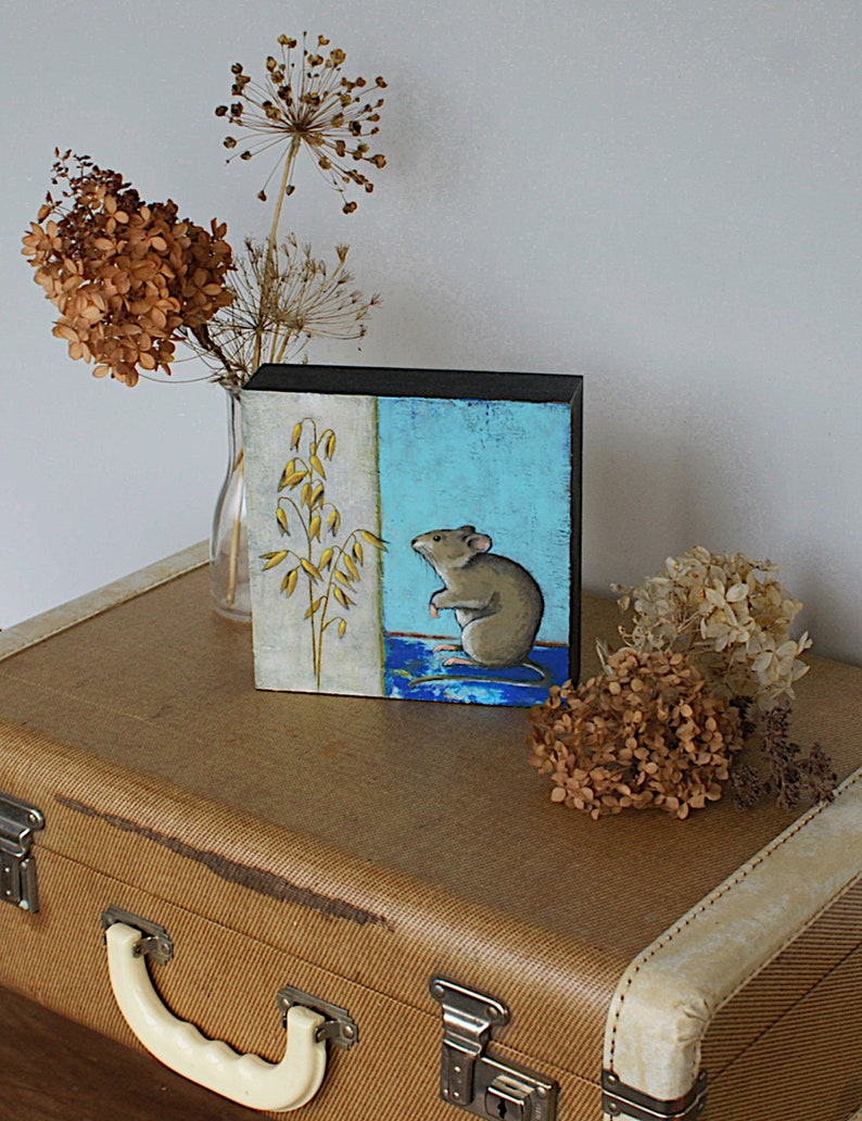 May include: A small painting on a wooden panel depicts a brown mouse standing on a blue and green background. The mouse is looking to the left, and there is a stalk of oats on the left side of the painting.