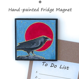May include: A square fridge magnet featuring a hand-painted black raven perched on a rock against a large red sun and a blue sky with small yellow stars. The text "Aimant pour frigo peint à la main" and "Hand-painted Fridge Magnet" appear above.