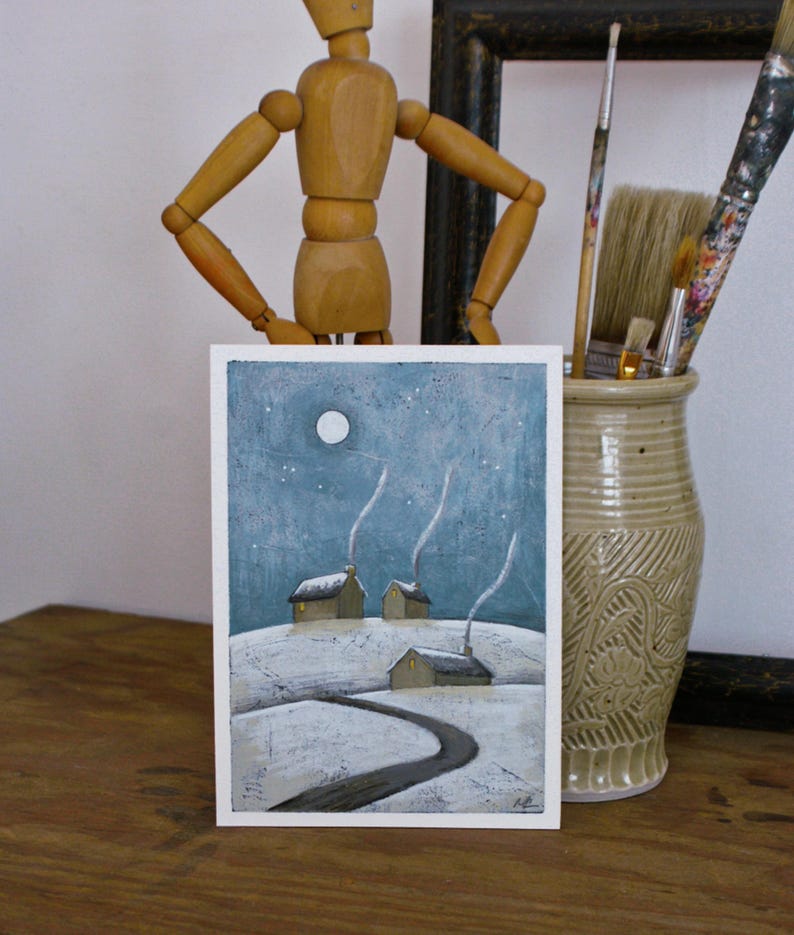 May include: A rectangular art print featuring a winter landscape with three small houses on a snow-covered hill under a blue night sky with a full moon and stars. A dark winding path leads toward the houses, each with a chimney emitting thin white smoke.