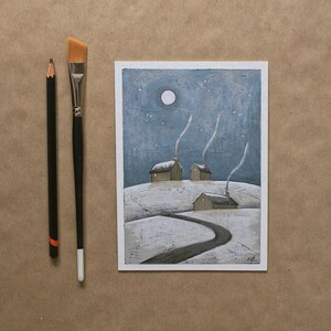 May include: A rectangular painting on paper depicting a snowy landscape with three small houses under a night sky with a full moon and stars. A black pencil and a flat-tipped paintbrush with a black handle are positioned to the left of the art.