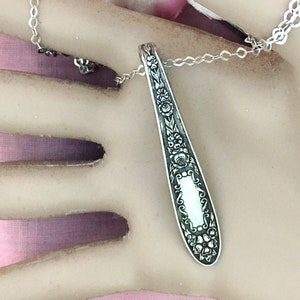 May include: A silver pendant necklace featuring a spoon-shaped design. The pendant is adorned with floral patterns and hangs from a delicate chain. The necklace is displayed on a neutral background.