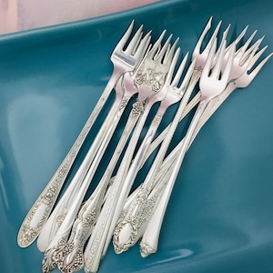 May include: A collection of antique silver forks with ornate floral and geometric patterns. The forks are arranged on a teal-colored surface, showcasing their intricate designs and polished finish. The tines are sharp and the handles feature detailed engravings.
