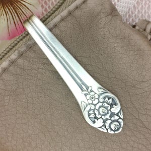 May include: A silver spoon with a floral design on the handle. The spoon is set against a neutral background. The handle features detailed carvings of flowers and leaves.