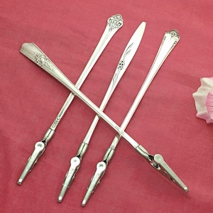 May include: Four silver-toned vintage serving pieces, possibly sugar tongs or similar, with decorative floral and geometric patterns on the handles. The pieces are arranged in a crisscross pattern against a red background.