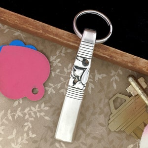 May include: A silver-colored key chain with a floral design and a key ring. The key chain has a rectangular shape with horizontal lines and floral engravings. The key chain is on a wooden tray with other keys and a pink tag.