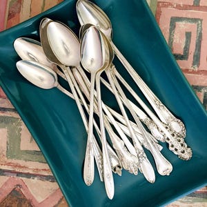May include: A collection of antique silver spoons arranged on a teal-colored rectangular tray. The spoons have various handle designs, some with ornate floral patterns. The spoons are reflective, and the tray sits on a patterned surface.