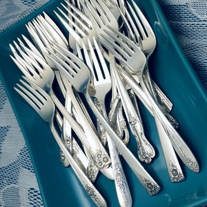 May include: A collection of silver forks with ornate floral and geometric handle designs, arranged in a teal-colored rectangular dish. The forks are made of shiny metal and are of varying sizes.