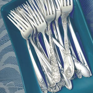 May include: A collection of ornate silver forks with detailed floral and grape designs. The forks are arranged in a turquoise rectangular dish, showcasing their polished surfaces and intricate patterns. The silverware is ideal for formal dining.
