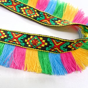 Op de afbeelding: Een zwarte en kleurrijke trim met franjes. De trim heeft een geometrisch patroon en is gemaakt van geweven stof. De franjes zijn in tinten roze, geel, groen en blauw.