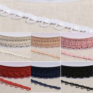 Pode incluir: Um close-up de dez rolos de guarnição elástica em várias cores, incluindo branco, bege, rosa, vermelho, azul marinho e preto. A guarnição apresenta um design de laço decorativo.