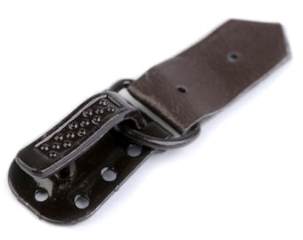Op de afbeelding: Een bruin leren band met een metalen gesp en een metalen lus. De gesp heeft een getextureerd oppervlak en een kleine metalen pen.