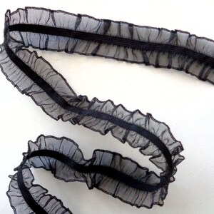 Puede incluir: Adorno elástico negro con borde fruncido. El adorno está hecho de una tela transparente y tiene una banda elástica negra que recorre el centro.