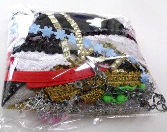 May include: A clear plastic bag filled with a variety of craft supplies, including lace, ribbon, chain, and beads. The supplies are in a variety of colors, including black, white, red, gold, green, and blue.