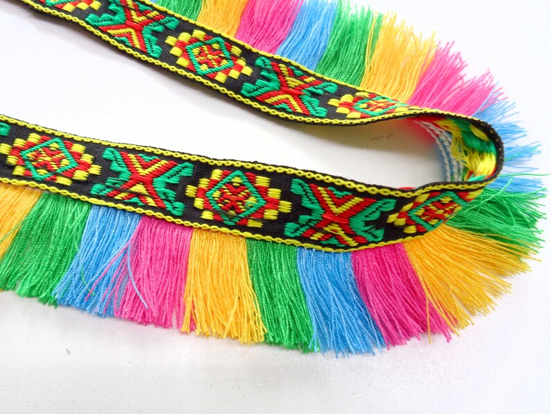 Op de afbeelding: Een zwarte en kleurrijke trim met franjes. De trim heeft een geometrisch patroon en is gemaakt van geweven stof. De franjes zijn in tinten roze, geel, groen en blauw.