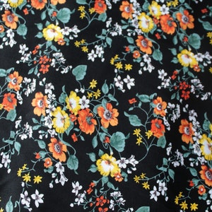 Op de afbeelding: Zwarte stof met een bloemenpatroon. Het ontwerp toont oranje, gele en witte bloemen met groene bladeren en stelen. De bloemen zijn verspreid over de donkere achtergrond.
